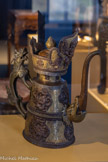 <center>Aiguière (hu), de type duomu</center>Fait au Népal, pour le Tibet XIXe siècle.
Cuivre, fer et bronze doré.
Cette aiguière de forme duomu, liée à la culture tibétaine lamaïque, témoigne d’influences multiples. La forme est dérivée du récipient tibétain bey-lep, « seau pour le beurre » qui pouvait également recevoir le lait ou le vin produit dans les monastères.
Les motifs sont d’inspiration chinoise et indienne. Des dragons lovés forment médaillons et paraissent en ronde-bosse sur le corps de l’objet orné d’arabesques gravées (patra), d’origine tibétaine. Son col rappelle la tiare jaune des moines lamaïstes tibétains. Un garuda, oiseau mythique issu du panthéon brahmanique, flanqué de deux makara, animal du bestiaire fantastique indien, en domine le sommet. La formule sacrée « om mani padme hüm » (le joyau dans le lotus) est inscrite sur le pourtour du col.
Le syncrétisme artistique de cette pièce s’explique certainement par les différents mouvements migratoires qui, du XVIIIe au XXe siècles, ont permis le transfert de savoirs et de techniques entre le Tibet, l’Inde et le Népal. De nombreuses pièces en métal ont ainsi pu transiter depuis Patan, dans la vallée de Kathmandu, jusqu’à Lhassa, par le biais de pèlerins ou de négociants.