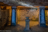 <center>Les Catacombes de Paris. </center> La lampe sépulcrale. Dans un renfoncement s'élève une colonne de pierre surmontée d'une vasque de forme antique, dite « lampe sépulcrale ». Ce monument, le premier réalisé dans l'ossuaire, servait à brûler de la résine de poix, l'air étant progressivement corrompu par les dépôts d'ossements, ce qui rendait l'air difficilement respirable pour les ouvriers chargés des transferts. L'entretien d'un foyer était en effet le meilleur moyen d'assurer une ventilation lors de travaux souterrains. Elle servait ainsi à veiller les morts et, plus prosaïquement, à améliorer la circulation de l'air, avant la construction des puits d'aération