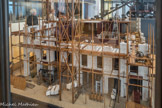<center>Musée des Arts et Métiers.</center> Immeuble en construction rue de Rivoli. Un chantier bien organisé.
Cette maquette très détaillée présente à la fois les outils et les techniques du bâtiment et illustre la nécessaire collaboration entre les artisans du gros œuvre : maître maçon, appareilleur, maçon limousinant.
Chaque matériau — pierre, plâtre, fers, briques — est stocké à un endroit précis du chantier et suit un chemin particulier ; les plus légers sont acheminés à l’aide d’un treuil à main et poursuivent leur route dans une brouette.
La locomobile, grâce à un système de courroies, anime les treuils des deux grandes sapines et fait tourner un malaxeur à mortier situé sous les arcades