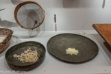 <center>Ensemble d’outils utilisés pour la récolte de la résine du lentisque appelée mastiha</center>11 Tamis [koskino], vers 1950. Bois, métal.
Les impuretés tombent et le mastiha plus propre reste dans le tamis.<br>
13 Plat [tapsi], vers 1900.
Cuivre.
Utilisé par une seule femme, il sert à contenir le mastiha en vrac avec les impuretés et à en faire le tri à la main : le mastiha plus propre d’un côté et les impuretés de l’autre.<br>
14 Grand plat [sini], vers 1900.
Cuivre.
Utilisé pour trier le mastiha des impuretés après la récolte.
On met au centre le tas de mastiha brut avec les impuretés et on met le mastiha trié vers les bords. Quatre femmes pouvaient travailler simultanément autour de ce grand plateau.
6B Terre Blanche, 2019
C’est sur cette terre pulvérulente que tombent les gouttes de résine.<br>
12 Outil  [timitiri], vers 1900
Fer, bois.
Sert à séparer la résine de mastiha de ses impuretés incrustés sur elle (terre, feuilles...).
