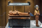 Figurine osirienne en bois de Toutânkhamon allongé sur une bière
Les différentes enveloppes funéraires et les bandelettes de la momie sculptée, ainsi que la tête et les pattes de lion du lit protègent le personnage contre les forces surnaturelles.
Bois, feuille d’or.
Coffret en bois en forme de sarcophage pour la figurine osirienne de Toutânkhamon avec couvercle ancré au coffret par des chevilles
Pour en assurer la protection, la momie de Toutânkhamon a été placée dans plusieurs cercueils qui furent ensuite enchâssés dans un lourd sarcophage en granite.
Bois, résine noire.