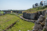 Le Fâ Barzan. <br> Le sanctuaire.
L'aire sacrée du sanctuaire est délimitée par un péribole qui la sépare de l'espace profane des rues périphériques. Le sanctuaire du Fâ couvre une surface rectangulaire de 12000 m2. La galerie à portique sud est longue de 119.40 m pour une largeur interne de 5.60 m, soit environ 670 m2. Les maçonneries ne sont connues qu'en fondation. Les murs sont construits avec :
• des appareils rectangulaires (18 cm sur 9 cm) en parement d'un noyau en béton, avec des joints marqués au fer ;
- des moellons sommairement équarris sur les parements non visibles.
L'intérieur de la galerie et de la cour est comblé par un remblai calcaire de 0 à 2,50 m de hauteur qui compense la pente de l'ancien sanctuaire et correspond au niveau de circulation de la nouvelle aire sacrée.
Ces constructions ont été reconstituées, à l'angle sud-ouest, sur la base des observations archéologiques. Un rang d'ardoises sépare les constructions antiques des reconstitutions.
Pour la partie ouest de cette galerie, partiellement connue en fouille, il a été choisi de représenter le péribole à l'aide de gabions posés à l'aplomb des vestiges. Les dés calcaires correspondent à l'emplacement des bases de colonnes de ce portique. La surface de pose était horizontale dans l'Antiquité. Les paliers formés par les gabions correspondent à la déclivité actuelle du site.