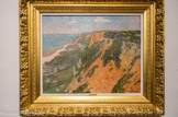 Les falaises de Quimper. MAXIME MAUFRA (1861 – 1918). Huile sur toile, 1905.
Élève du paysagiste Charles Leroux, Maxime Maufra s’initie à la peinture en plein air avec les frères Charles (1831 - 1911) et Alfred Leduc. En 1899, il abandonne le métier auquel le destine son père et se consacre exclusivement à la peinture. Maufra expose à la deuxième exposition des peintres impressionnistes et symbolistes en 1892 avec les peintres les plus avant-gardistes de son temps. En quittant la Bretagne, Maufra s’installe au Bateau-Lavoir à Montmartre.
Maufra montre ici qu’il est un virtuose du paysage. Ce peintre de la nature équilibre son tableau avec des couleurs chaudes d’été. La place majeure de la toile est occupée par la falaise rouge que quelques végétaux parsèment çà et là. En contrebas, proche de l’eau, une étendue verte légèrement cernée de noir rappelle que le peintre a parfaitement assimilé les enseignements de l’école de Pont-Aven. Le plan d’eau se distingue tandis qu’à l’horizon on aperçoit le rivage. Comme dans la plupart de ses œuvres, et fidèle à la leçon synthétiste, Maufra place très haut le ciel et lui accorde une place minime : une petite bande bleue traversée par quelques nuages gris. Ce ne sont pas des effets atmosphériques qui l’interpellent, mais la nature dans toute sa force