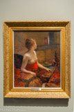 L’attente. FERNAND CORMON (1847 – 1925). Huile sur toile.
Élève de Portaels à Bruxelles, puis de Cabanel et de Fromentin à Paris, Fernand Cormon expose pour la première fois au salon de Paris en 1863. Adepte d’une peinture à sensation et certainement influencé par l’orientalisme de son maître Fromentin, le peintre a livré de nombreuses toiles d’inspiration orientale, telle La favorite déchue 1870 ou Jalousie au sérail 1874.
Une jeune femme, cheveux relevés et seins dénudés, est assise sur un sofa dont le tissu est à motifs orientaux. La pièce dans laquelle elle se tient est sombre et confinée. Quelques objets évoquent l’oisiveté qui n’est d’ailleurs pas sans rappeler l’existence des femmes de la bonne société occidentale à la même époque : aiguières sur un plateau, mais surtout la fumée, matérialisée au travers du narguilé qui repose sur une table au premier plan. Comme si quelqu’un était sur le point de pénétrer dans la pièce, la jeune femme détourne son regard vers le fond de la pièce, de sorte que nous ne voyons pas son visage. Qu’attend la jeune esclave ? Est-elle sur le point de recevoir le sultan ? En peignant un « ailleurs », Fernand Cormon n’hésite pas à se livrer à une œuvre explicitement érotique dans laquelle la présence de l’homme, du séducteur, est perçue et non montrée, le spectateur du tableau prenant ainsi sa place.