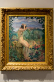 Nu à la fontaine. LEBASQUE (1865-1937). Huile sur toile, 1911
Henri Lebasque n'ignore rien de l’avant-garde de son temps. Comme les « poulains » du marchand Ambroise Vollard (1866-1939), il fait dès 1906 des essais de céramique chez André Metthey (1871-1920) à Asnières où il décore des vases et des plats.
D'abord paysagiste, Lebasque est particulièrement connu pour son travail sur les nus et sur la couleur. Lebasque regroupe ici deux thèmes qui lui sont chers dans ce que le critique d'art Adolphe Tabarant (1863-1950) appelle un « paysage animé » qui n'est ni véritablement un nu, et pas davantage un paysage. Comme il le remarque justement, Lebasque renouvelle le thème de la jeune fille gracile. Ici nue et de profil, les bras relevés, la figure occupe la place centrale de la composition. Elle se recoiffe, la tête penchée. Son bras dissimule son visage, totalement absent. Son chapeau repose à ses côtés. A l'arrière-plan, on distingue dans des camaïeux de bleus quelques nuages au-dessus de montagnes qui surplombent un plan d'eau tranquille. La scène semble totalement irréelle. Seule une silhouette de femme portant une robe rose sur le chemin de terre ancre la composition dans la réalité.